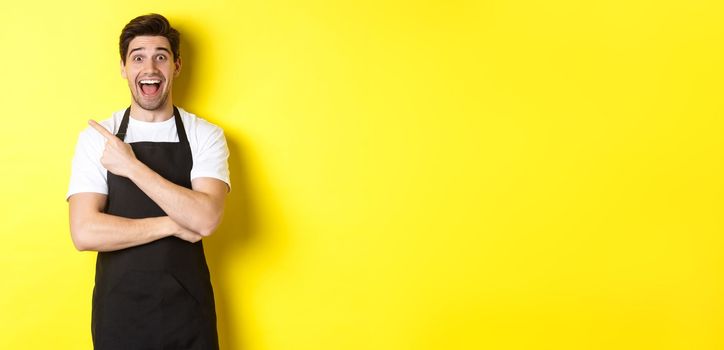 Surprised salesman in black apron pointing finger upper left corner, showing shop promo offer, standing against yellow background.