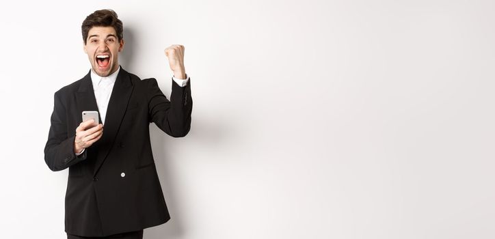 Portrait of happy handsome man in suit, rejoicing, achieve goal on mobile app, raising fist up and shouting yes, holding smartphone, standing against white background.