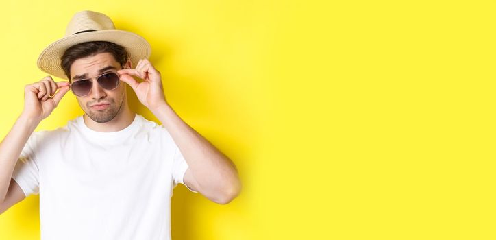 Concept of tourism and vacation. Close-up of cool tourist enjoying holidays on trip, wearing sunglasses with straw hat, yellow background.