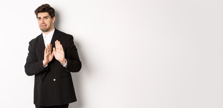 Image of stylish handsome man in black suit, cringe from something embarrassing, declining offer and showing stop gesture, standing against white background.