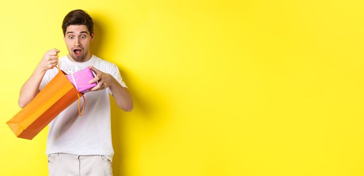 Concept of holidays and celebration. Young man looking surprised as take out gift from shopping bag, standing over yellow background.