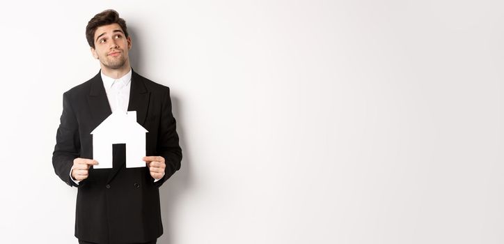 Image of handsome businessman in black suit, looking for home, holding house maket and gazing dreamy at upper right corner, standing against white background.