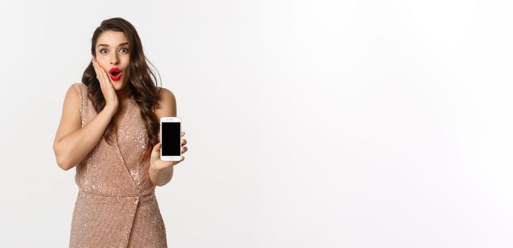 Online shopping. Elegant woman in luxury dress showing smartphone screen, looking amazed at camera, white background.