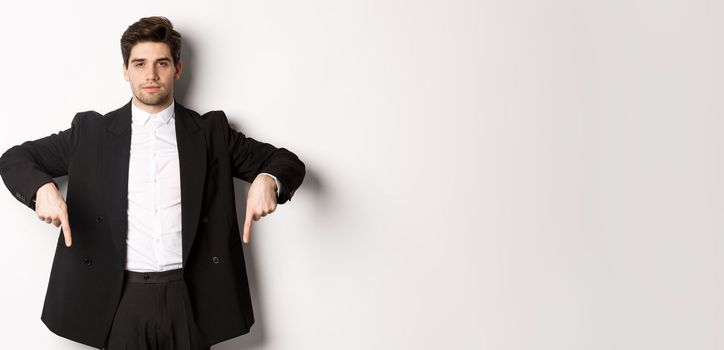 Image of handsome man dressed for formal party, wearing suit and pointing fingers down, showing advertisement or making announcement, standing over white background.