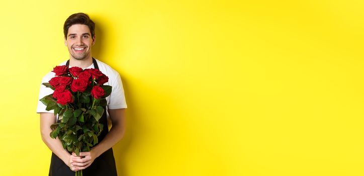 Smiling florist in black apron holding flowers, selling bouquet of roses, standing over yellow background.