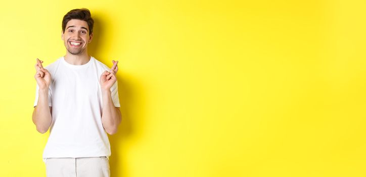 Hopeful smiling man holding fingers crossed, making wish or praying, standing over yellow background.