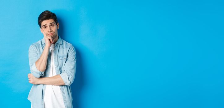 Unamused and bored man looking without interest at camera, standing against bllue background.