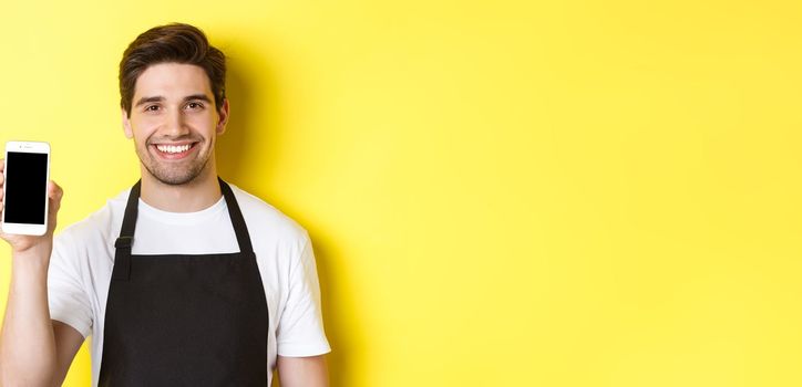 Close-up of handsome waiter in black apron showing smartphone screen, recommending app, standing over yellow background.