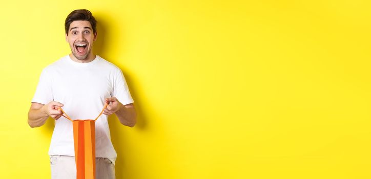 Surprised guy open shopping bag with fist, looking excited and happy at camera, standing against yellow background.