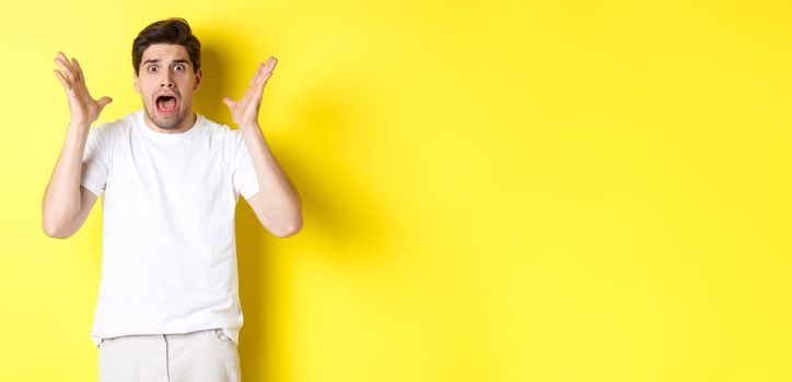 Frustrated and shocked guy panicking, screaming and looking scared, standing in white t-shirt over yellow background.