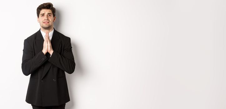 Image of handsome businessman in suit need help, begging you, holding hands in plead gesture and looking desperate, standing over white background.