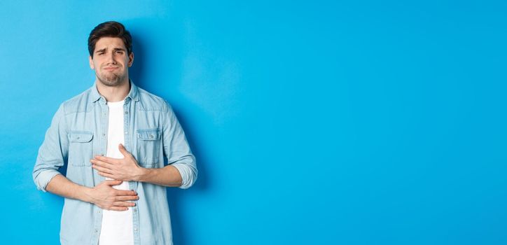 Man holding hands on belly and grimacing from pain, complaining on stomach ache, standing against blue background.