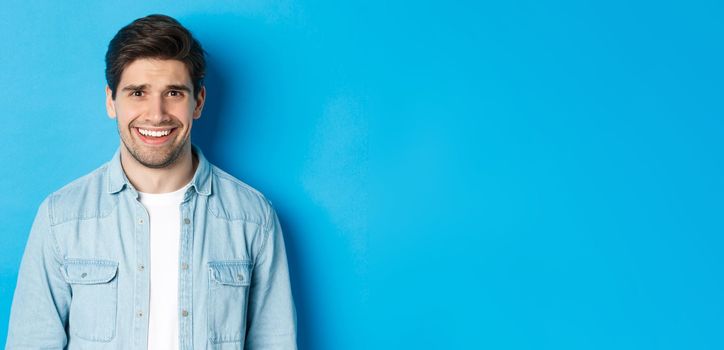 Close-up of young man feeling awkward, smile and cringe from uncomfortable situation, standing over blue background.