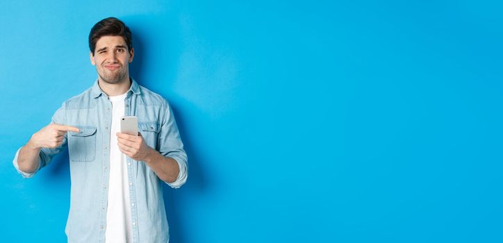 Concept of online shopping, applications and technology. Skeptical and displeased guy pointing finger at smartphone and grimacing disappointed, standing over blue background.