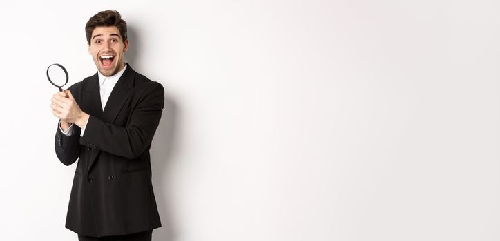 Handsome businessman in black suit, holding magnifying glass and smiling, found something, standing against white background.