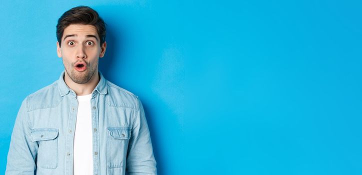 Close-up of handsome surprised man, open mouth and saying wow with amazement, hear announcement, standing over blue background.