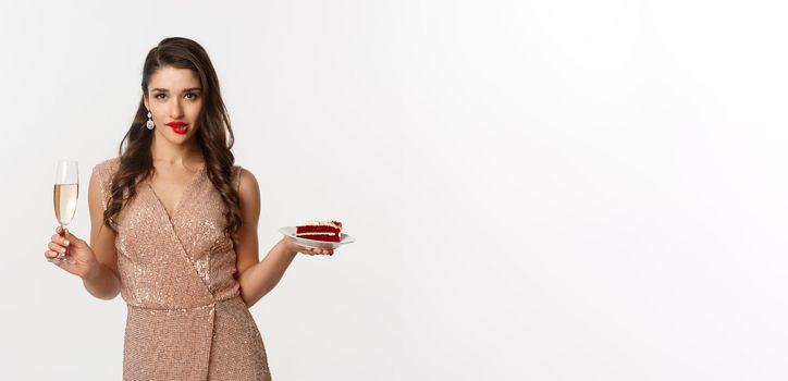 Party and celebration concept. Sexy woman in elegant dress, holding champagne and piece of cake, biting lip and looking seductive at camera.