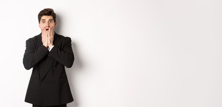 Image of concerned and scared man in black suit, gasping and looking alarmed at camera, standing over white background.