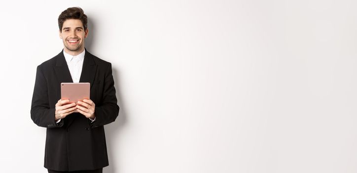 Image of handsome businessman in trendy suit, holding digital tablet and smiling, standing against white background.