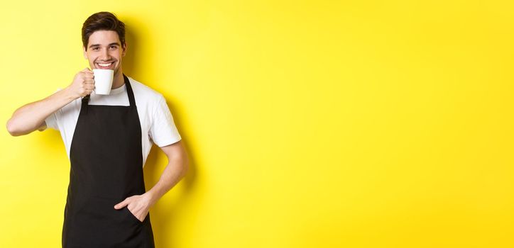 Cafe barista drinking cup of coffee and smiling, wearing black apron, standing over yellow background.