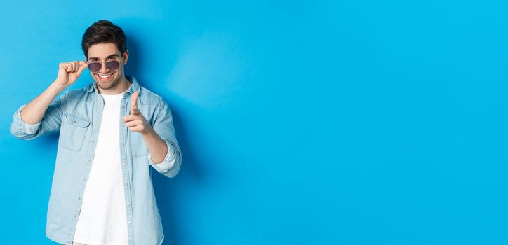 Handsome cheeky guy in sunglasses, smiling and pointing finger gun at camera, flirting with you, standing over blue background.