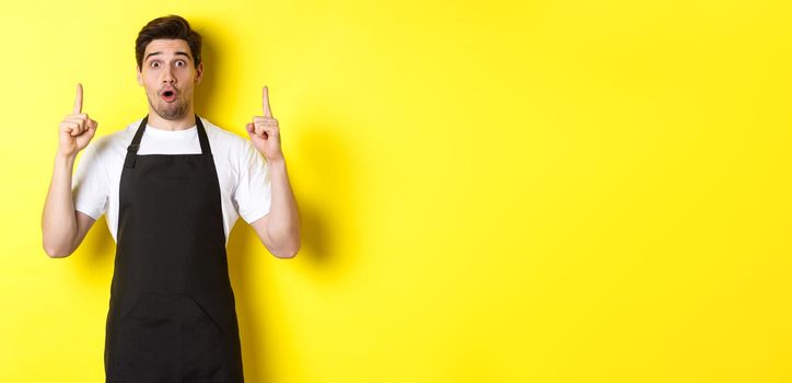 Surprised male waiter in black apron showing logo or advertisement, pointing fingers up at copy space, standing over yellow background.