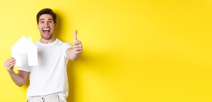 Real estate concept. Happy young male buyer showing thumb up and paper house model, smiling satisfied, standing over yellow background.