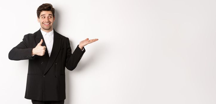 Handsome businessman in black suit, showing thumb-up and holding your product in hand over white copy space, standing against white background.