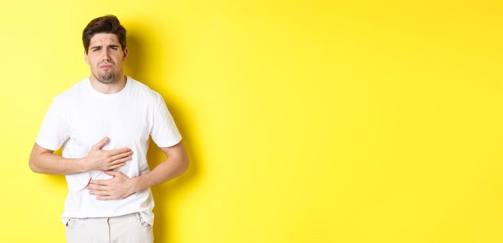 Man having stomach ache, grimacing from pain and touching belly, standing over yellow background.