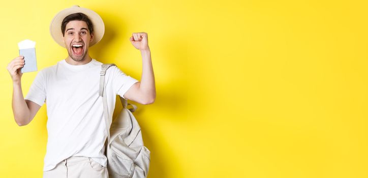 Tourism and vacation. Man feeling happy about summer trip, holding passport with plane tickets and backpack, raising hands up in celebration gesture, yellow background.