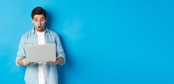 Impressed caucasian guy looking at laptop screen with amazement, checking out promo in internet, standing against blue background.