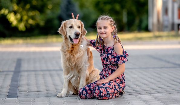 Preteen girl with golden retriever dog sitting in park in beautiful summer day. Adorable female child kid hugging purebred doggy pet outdoors portrait