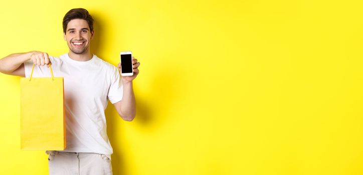 Concept of mobile banking and cashback. Young happy guy holding shopping bag and showing smartphone screen, yellow background.