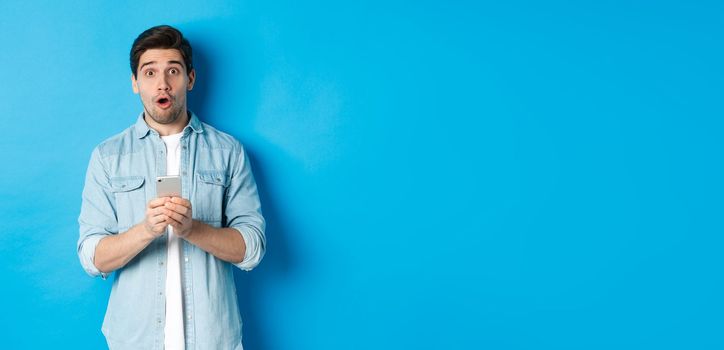 Surprised man in casual clothes looking astounded, holding smartphone, standing against blue background.