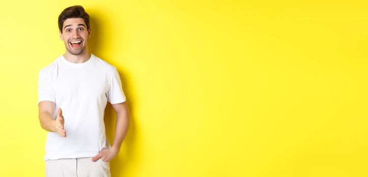 Friendly man greeting you with handshake, smiling amused, saying hello, standing over yellow background.