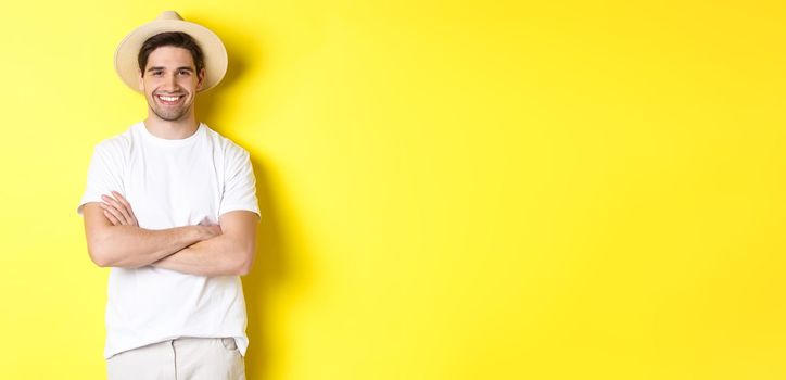 Tourism. Handsome young man looking happy, wearing straw hat for travelling, cross hands on chest and smiling, standing over yellow background.