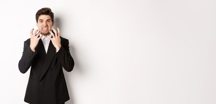 Image of angry businessman in suit, looking with furious expression and clenching fists, express hatred, standing mad over white background.