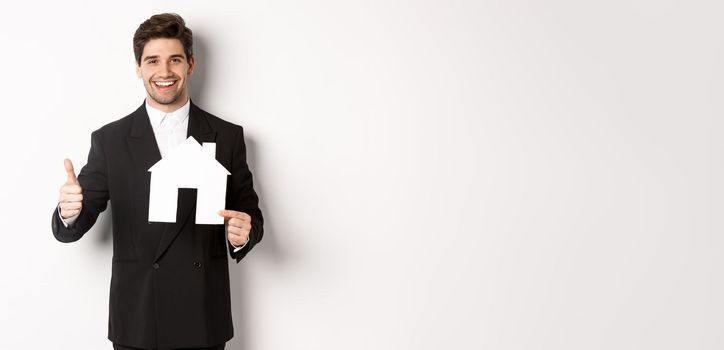 Portrait of confident real estate agent showing house maket and thumb-up, standing against white background.