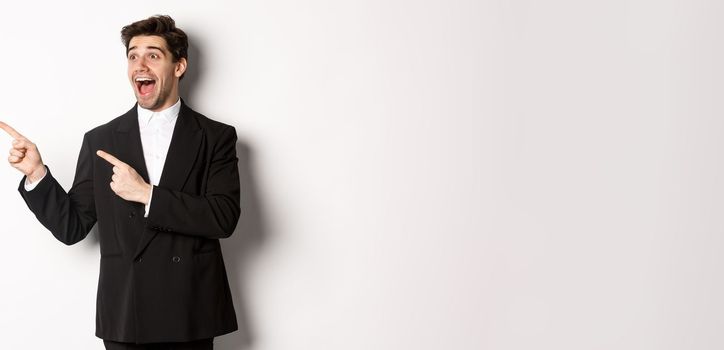 Portrait of amazed and happy handsome man in black suit, pointing and looking left impressed, showing awesome christmas promo, standing over white background.