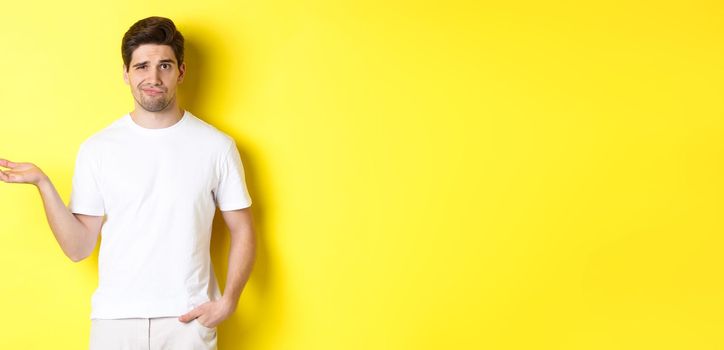 Confused and displeased guy raise hand, grimacing puzzled, standing against yellow background. Copy space