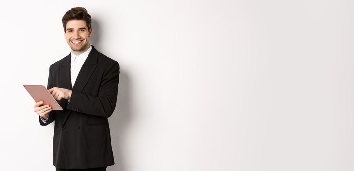 Image of handsome businessman in black suit, using digital tablet and smiling, standing against white background.