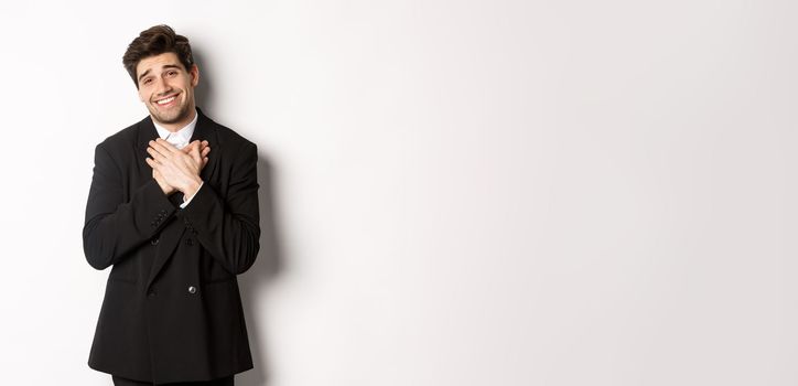 Portrait of happy and pleased handsome man in party suit, holding hands on heart and sighing flattered, saying thank you, standing grateful over white background.