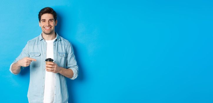 Smiling happy man pointing at paper cup with coffee, recommending cafe, standing over blue background.