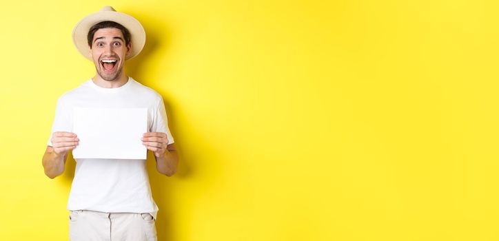 Excited tourist showing your logo or sign on blank piece of paper, smiling amazed, standing against yellow background.