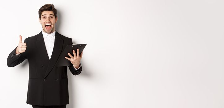 Portrait of confident businessman in black suit, holding clipboard with documents and showing thumb-up in approval, praise good job, standing against white background.