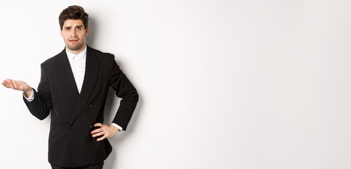 Image of confused handsome man in formal suit, raising hand and shrugging, cant understand something, standing against white background.