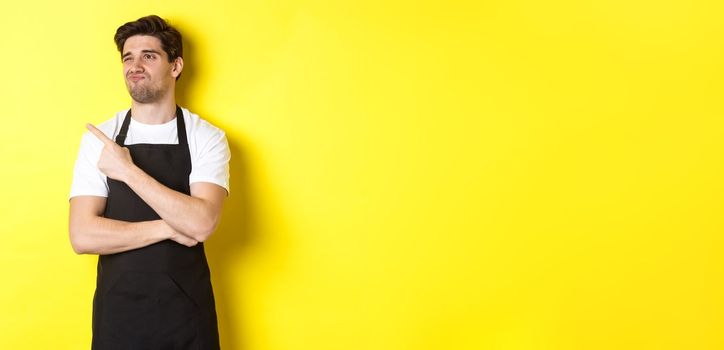 Skeptical male seller in black apron looking displeased, grimacing and pointing left at advertisement, standing over yellow background.