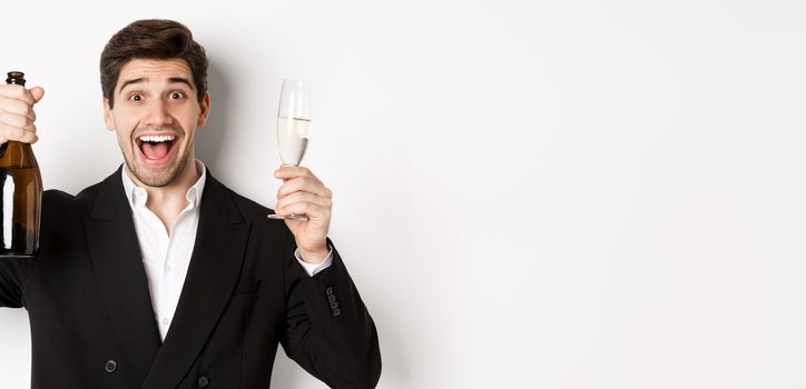 Close-up of handsome smiling man in black suit, making a toast, holding champagne and glass, celebrating new year, standing against white background.