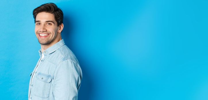 Handsome and confident guy turn head at camera, smiling happy, standing over blue background.