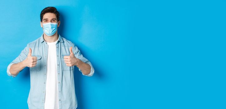 Concept of covid-19, pandemic and social distancing. Young man in medical mask showing thumbs-up in approval, like and agree, standing against blue background.